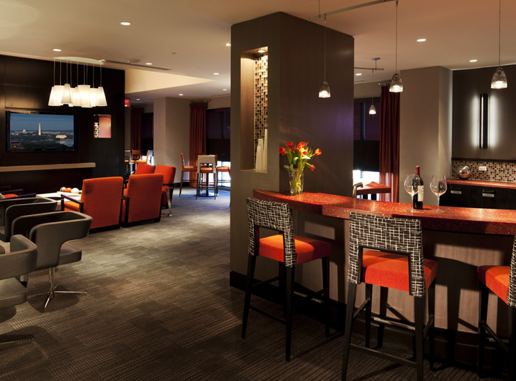 Clubroom kitchen. Bar area with four bar stools overlooking kitchen with tv on wall. Orange and wood accent colors with artwork on wall. Additional seating in background, 4 top table and orange accent chairs next to tv